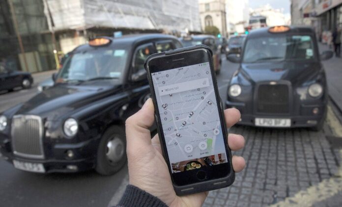 Uber app on a mobile telephone, as it is held up for a posed photograph, with London Taxis in the background, in London