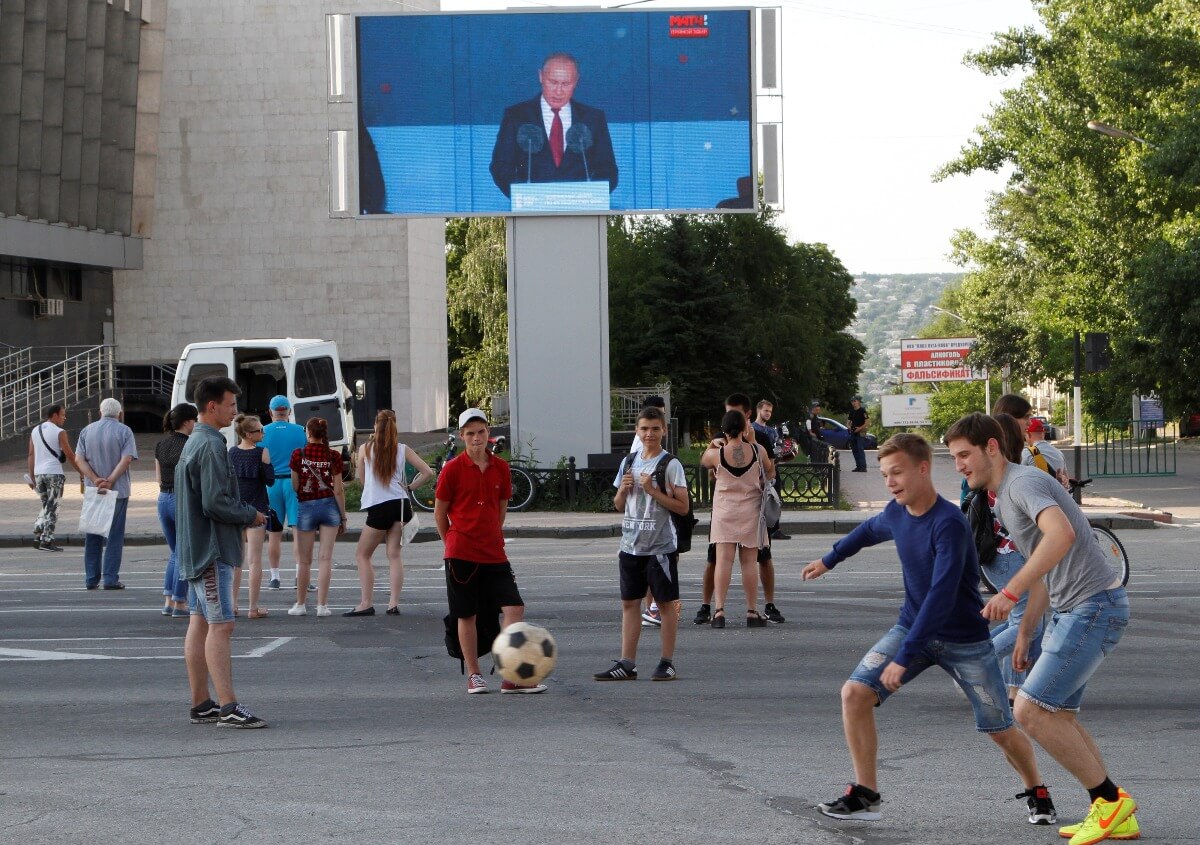 Youths play with a ball as Russian President Vladimir Putin is seen on a screen delivering a speech before the opening match